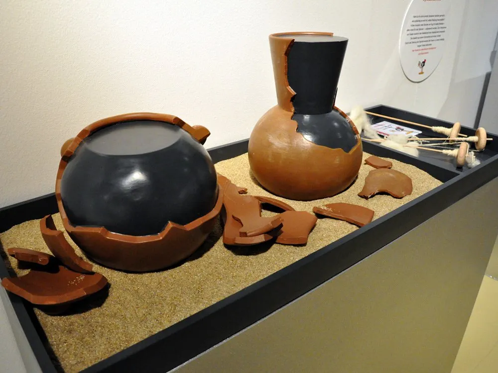 Interaktivstation: zwei beschädigten Keramikgefäßen auf Sand, umgeben von Magnet-Scherben zum Anbringen