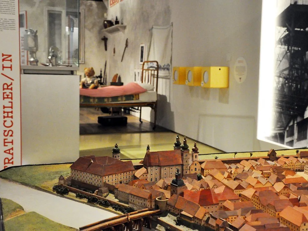 Ausstellungsraum: Modellstadt mit roten Dächern und einer Kirche, im Hintergrund ein Bett mit rosa Decke und eine Vitrine mit "TRAISCHER/IN"-Schild