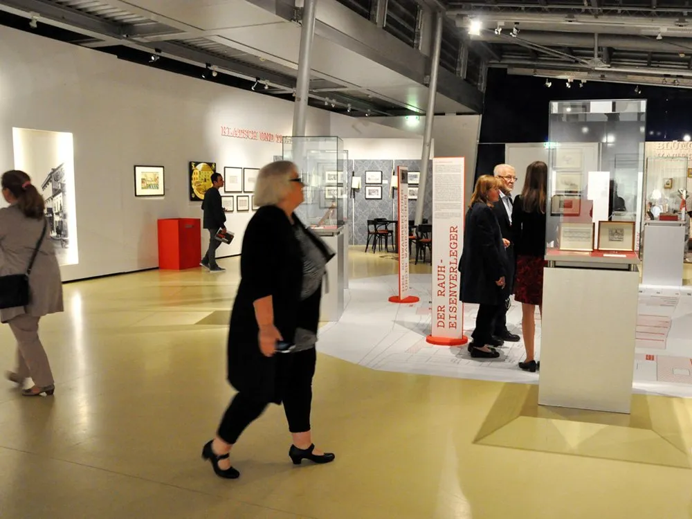 Ausstellungsraum: Besucher sehen Fotos und Schilder wie "DER EISENVERLEGER". Eine Karte liegt am Boden, historische Bilder hängen an den Wänden