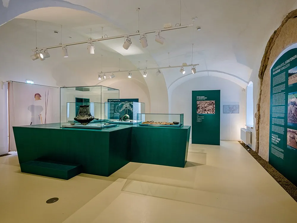 Ausstellung mit grünen Vitrinen mit antiken Gefäßen und Artefakten. Wände mit Infopanelen