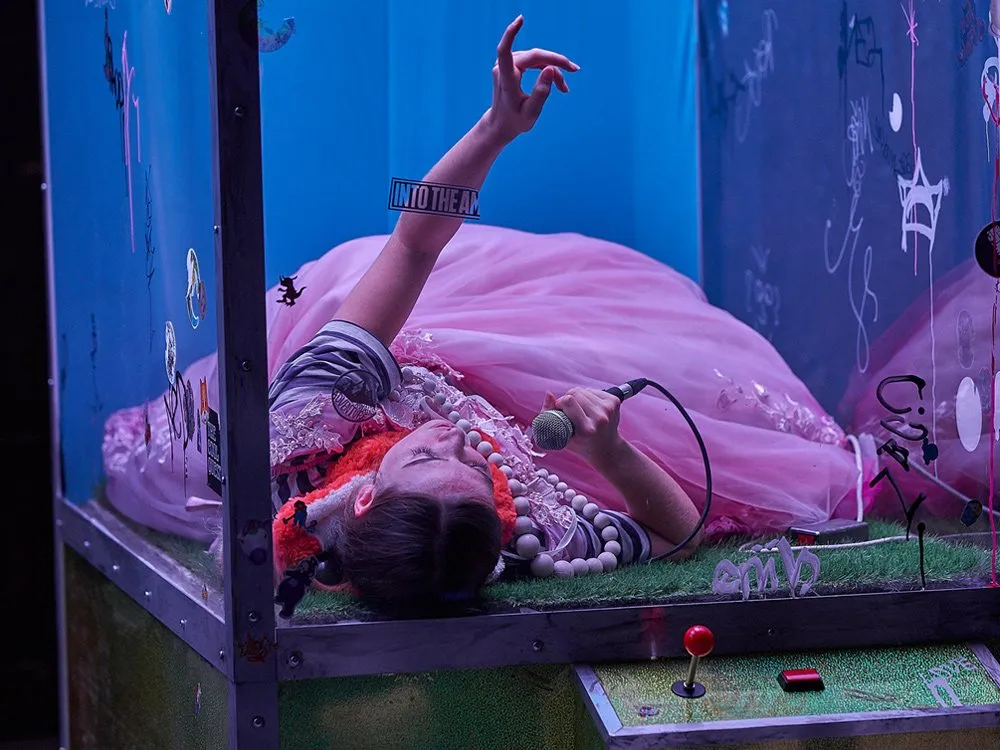 Person in einem rosa Kleid liegt in einer Glasvitrine mit grünem Boden, umgeben von bunten Accessoires und Mikrofon