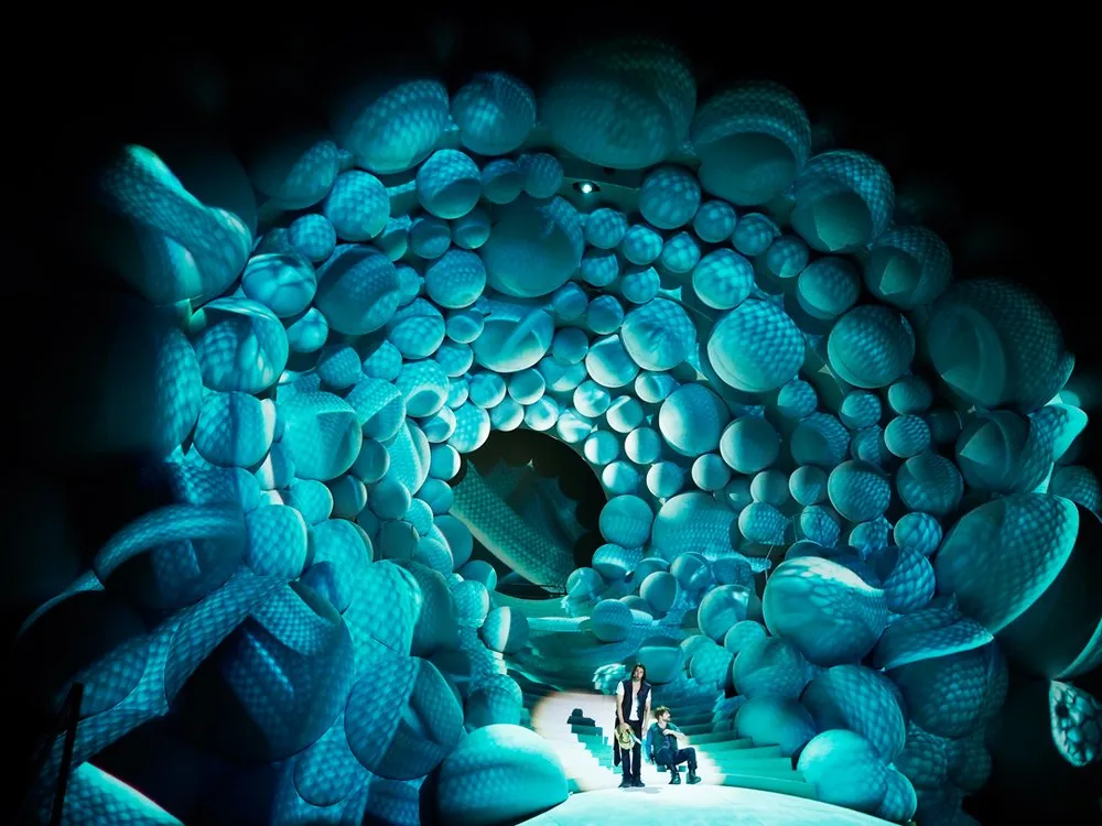 Szene: Drei Personen in einem Tunnel aus blauen, kugelförmigen Strukturen, die einen himmlischen Wolkenstrudel darstellen