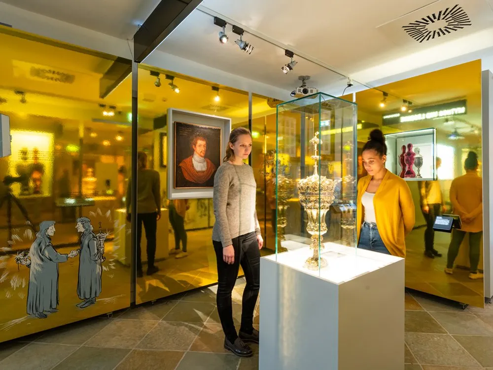 Ausstellungsraum mit Glaswänden, Gemälde, Zeichnungen und einer Vitrine mit goldenem Kunstobjekt, betrachtet von zwei Personen.