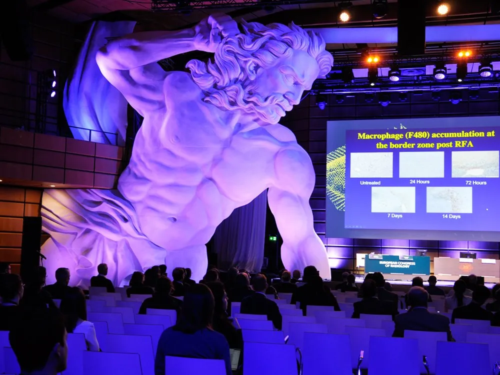 Riesiger Poseidon-Statue, Publikum auf Stühlen und einer Präsentation auf Leinwand.