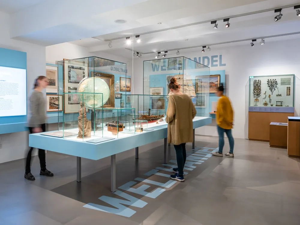 Ausstellungsraum: Vitrinen mit antiker Weltkugel, Schiffsmodell und Statuen, umgeben von Bildern und Besuchern, Schriftzug „WELTHANDEL“ auf Boden