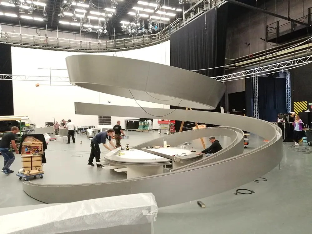 Arbeiter bauen eine Bühne mit geschwungenen Elementen, umgeben von Studiotechnik und Materialien