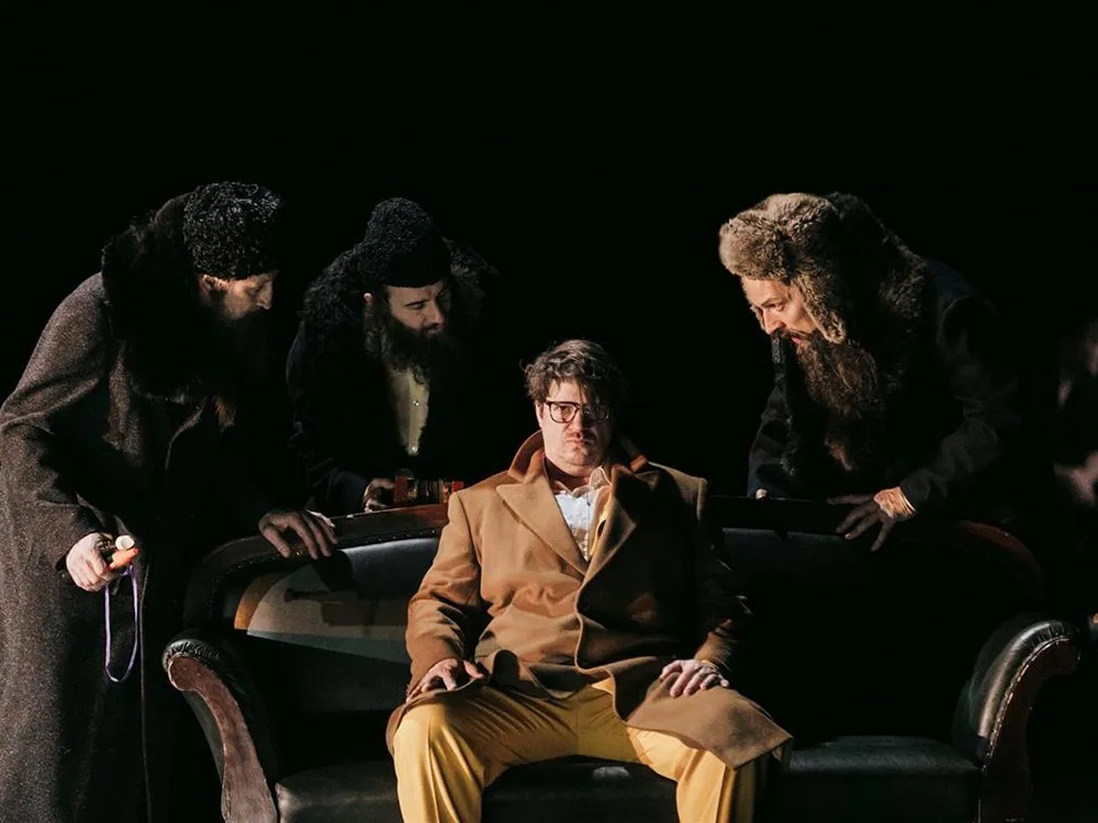 Schauspieler in braunem Mantel und gelben Hosen sitzt auf einem Sofa, umgeben von drei Figuren in Pelzmänteln