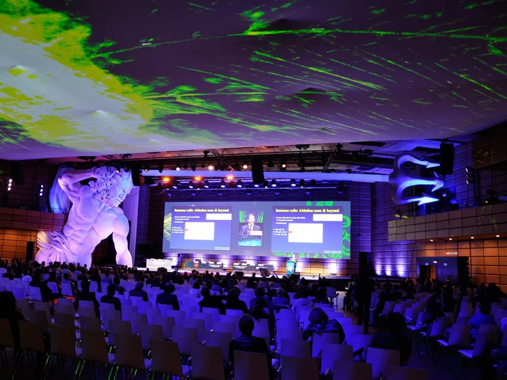 Beleuchteter Konferenzsaal mit großer Poseidon-Statue, Publikum auf Stühlen und zwei Leinwände mit Präsentationen zu "Immune cell Ablation" und "Beyond"