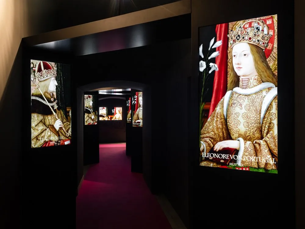 Ausstellungsraum: Dunkler Gang mit rotem Teppich, gesäumt von beleuchteten Porträts von Friedrich III. und Eleonore von Portugal