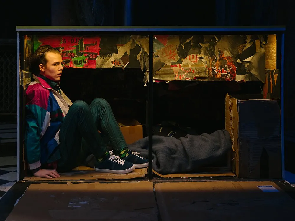 Schauspieler in bunter Jacke sitzt in einem kartonartigen, mit Plakaten und Decken dekorierten Bühnenbild