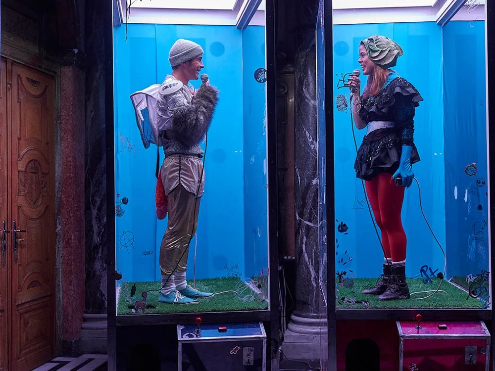 Zwei Schauspieler in Glasvitrinen sehen sich an und halten Mikrofone