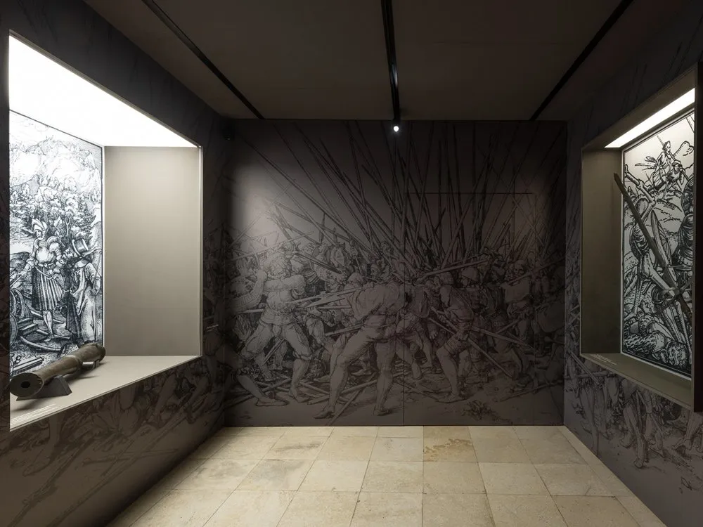 Ausstellungsraum: Dunkler Raum mit gefliestem Boden, bemalte Schlachtszenen an den Wänden und einer Kanone in einer beleuchteten Nische