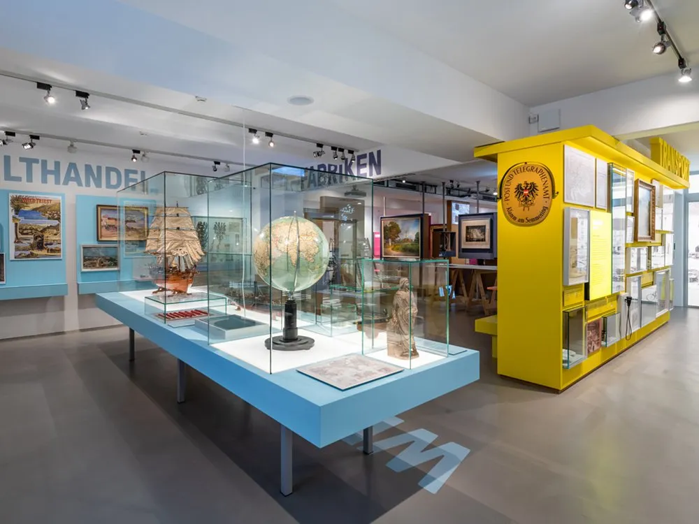 Ausstellungsraum: Vitrine mit einer Weltkugel, Schiffsmodell und Statuen, umgeben von Bildern und einem gelben Schrank, mit dem Schriftzug „WELTHANDEL“ und „FABRIKEN“ an den Wänden