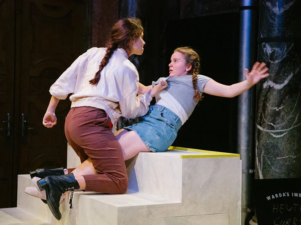 Zwei Schauspielerinnen auf weißen Treppen, eine packt die andere am Shirt