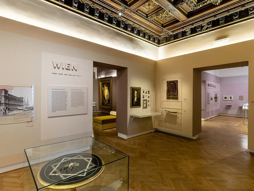 Ausstellungsraum: verziertes Deckenfries, Holzboden, historischen Gemälden, Fotografien und einer Vitrine