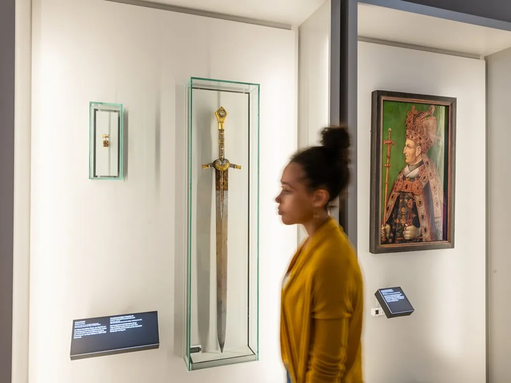 Ausstellungsraum: Vitrine mit einem Schwert und kleinere Artefakten, neben Gemälde eines Herrschers, betrachtet von einer Person