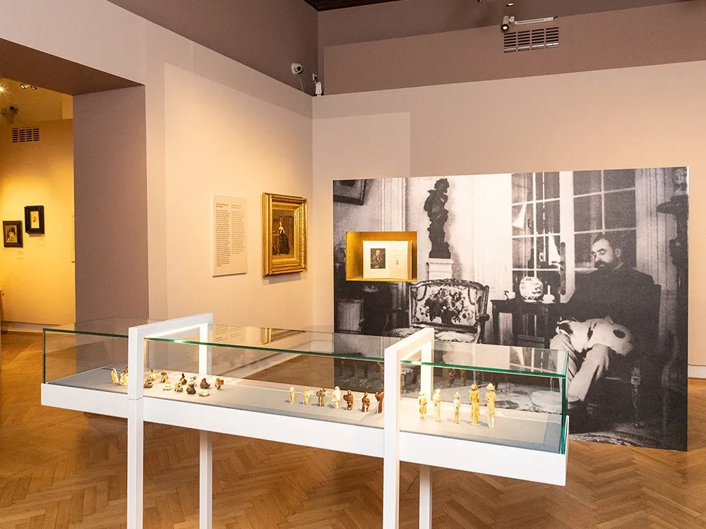 Ausstellungsraum: Glasvitrinen mit kleinen Skulpturen, historischen Gemälden an den Wänden und einem großen Foto eines sitzenden Mannes mit Skulptur im Hintergrund