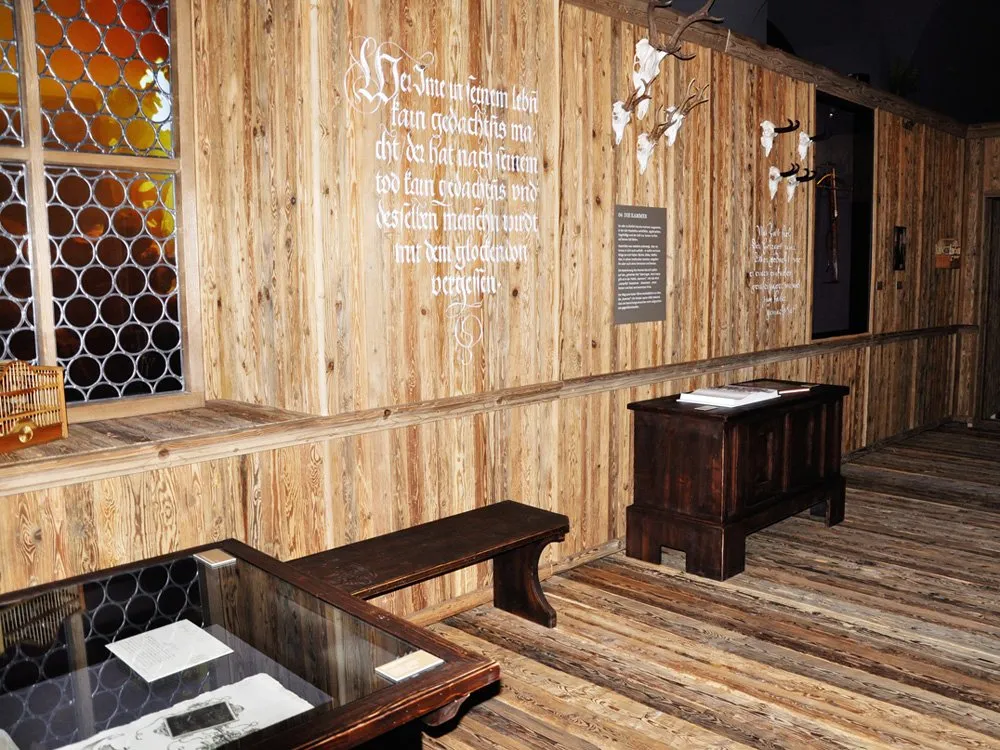 Rustikaler Raum mit Holzwänden und -boden, Geweihen, antiken Möbeln, buntem Glasfenster und Vitrine mit Dokumenten.