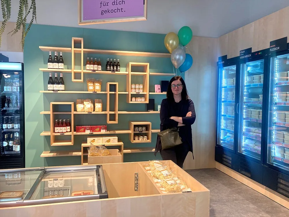 Frau vor einer Holztheke mit gefrorenen Lebensmitteln, umgeben von Kühlregalen, hängenden Pflanzen und Luftballons, sowie einem Schild mit dem Text „für dich gekocht“.