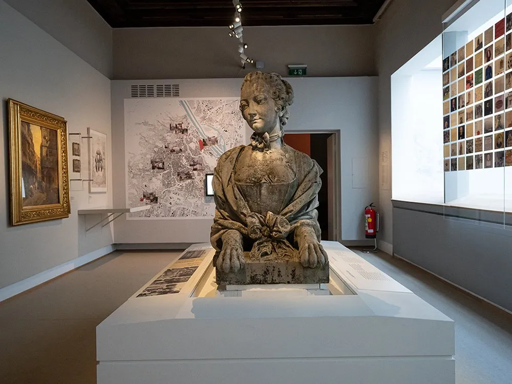 Ausstellungsraum: Alte Statue einer Frau in historischer Kleidung auf einem weißen Sockel, umgeben von Gemälden, einer detaillierten Stadtmappe und einer Wand mit vielen kleinen Fotos.