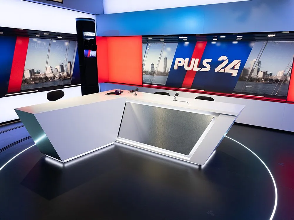 Weißes, futuristisches L-förmiges Pult mit Mikrofonen und Stühlen, umgeben von großen Bildschirmen mit Stadtansicht und dem Logo "PULS 24" in Blau und Rot