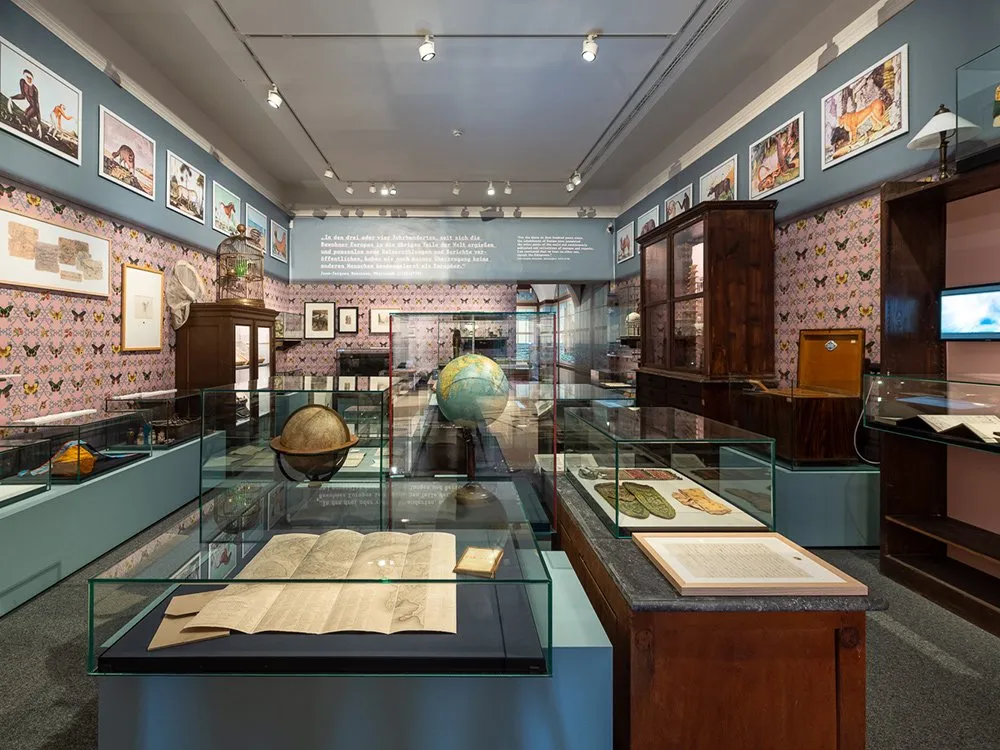 Ausstellungsraum: Vitrinen zeigen historische Karten, Bücher und Artefakte, während an den Wänden Gemälde und Fotos mit Szenen von Reisen und exotischen Landschaften sowie mehrere Globusse ausgestellt sind.