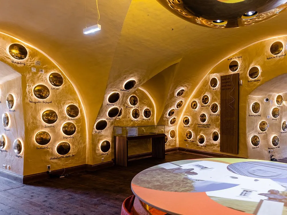 Gewölbter Raum mit gelb erleuchteten Wänden, dekoriert mit runden, beleuchteten Bildern und Beschriftungen verschiedener Orte, sowie einer Vitrine und einem runden Tisch mit Projektionen.