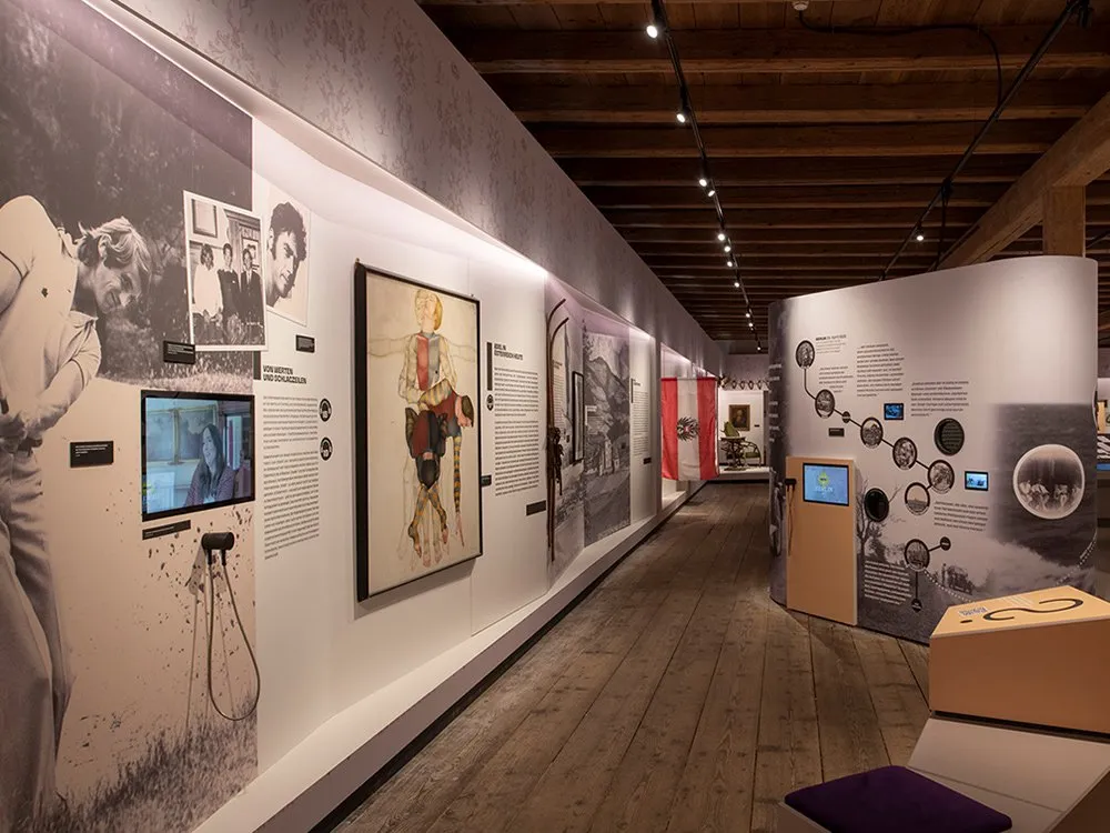 Ausstellungsraum: Langer Saal mit Wänden, die historische Fotos, ein Gemälde mit Mann in traditioneller Kleidung, Infotafeln sowie einen Monitor und interaktive Stationen zeigen.