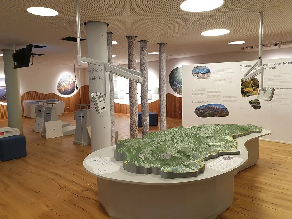 Ausstellungsraum: Landschaftsmodell eines bergigen Gebietes auf einem Tisch, Projektor hängt über dem Modell, im Hintergrund sind Plakate mit Text und Bildern.