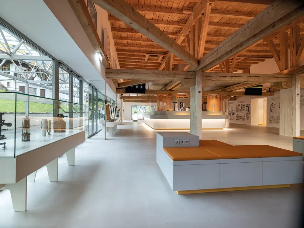 Heller Ausstellungsraum mit hoher Holzbalkendecke, großen Fenstern und Vitrinen