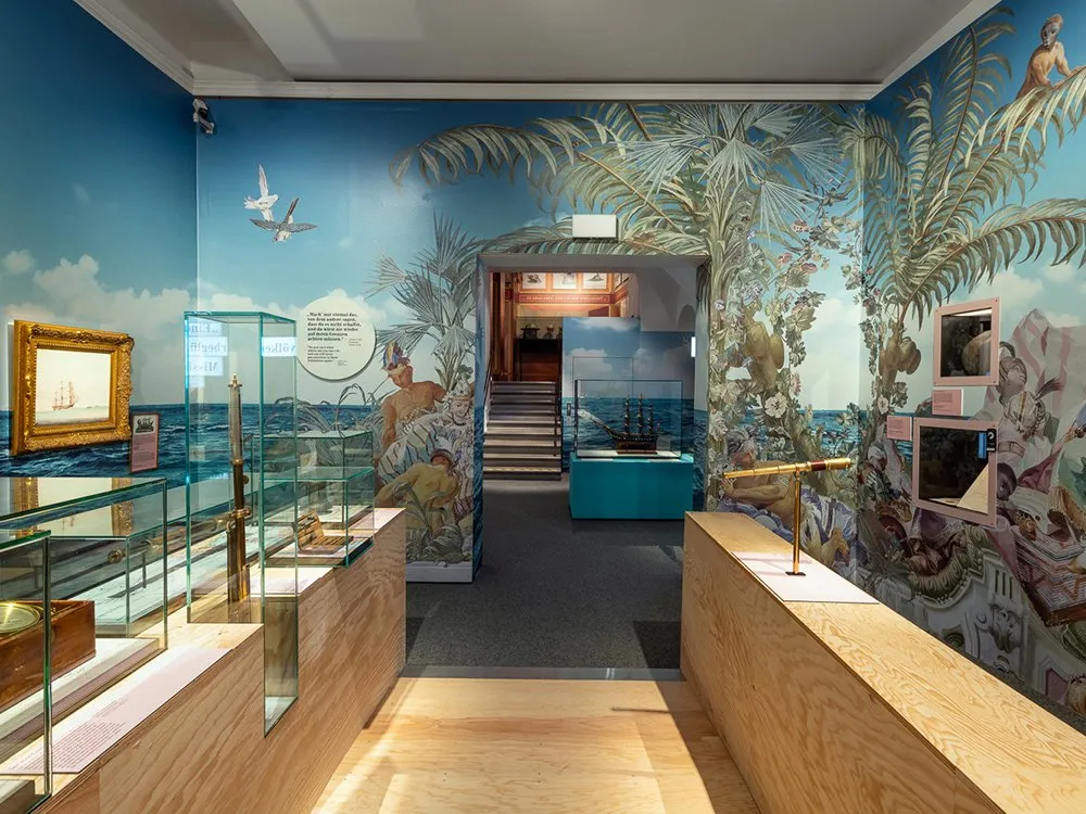 Ausstellungsraum: Wände mit tropischen Wandmalereien und historischen Gemälden, darunter ein Bild eines Segelschiffs. Vitrinen zeigen nautische Instrumente und ein Modell eines Schiffes, ergänzt durch eine Teleskopstation.
