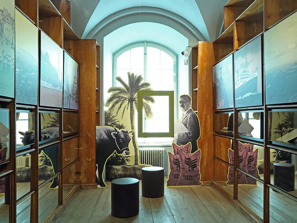 Ausstellungsraum: Schwarz-Weiß-Foto eines Mannes in einem Anzug vor einer Palme, daneben ein Kuh- und Kaffeesack-Motiv, umgeben von Holzregalen mit Bildern von Schiffen und Städten