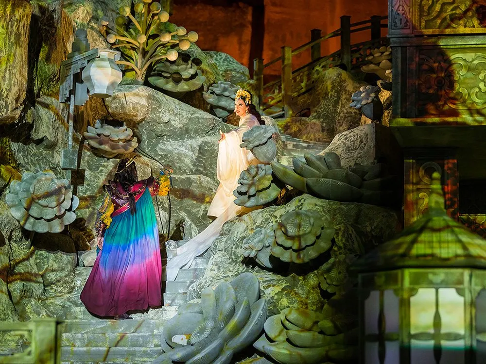 Szene: Zwei Figuren in farbenfrohen Gewändern auf einer steinigen Treppe mit großen Blättern, beleuchtet von einer Laterne, vor einem dekorativen Hintergrund mit Brücke.