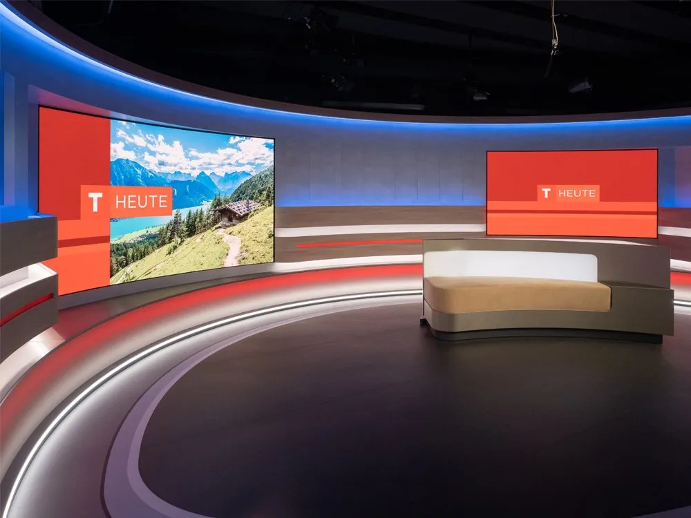 "Tirol heute"-Studio mit gebogener Bühne, LED-Wänden mit Berglandschaft, rotfarbenen Akzenten und einem zentralen Pult