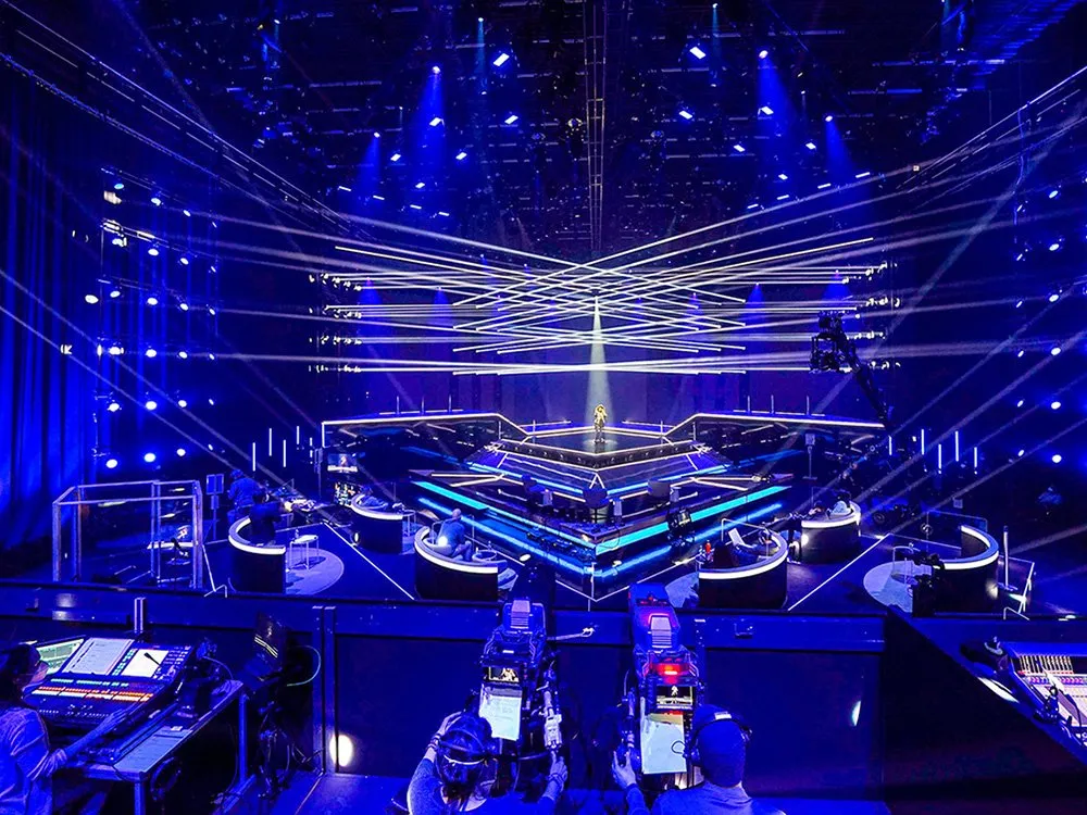 Futuristische Bühne mit blauen Lichtern und Laserstrahlen, die sich in der Mitte treffen. Die mehrstufige Plattform mit leuchtenden Kanten ist von Technik und Kameras umgeben.