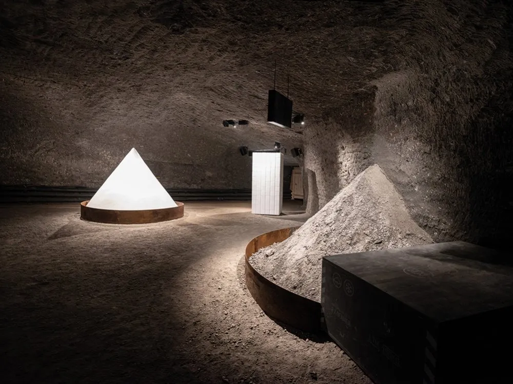 Unterirdische Ausstellung mit rauer Felsdecke, leuchtender weißer Pyramidenkegel, einem Haufen Salz und einer weißen Vitrine unter moderner Beleuchtung.