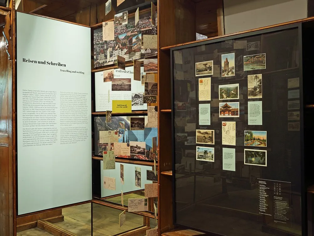 Ausstellungsraum: Holzregale mit Postkarten und Fotos, darunter Reise- und Schreibthemen von Stefan Zweig, mit Texttafeln und einem Spiegel an der Seite.