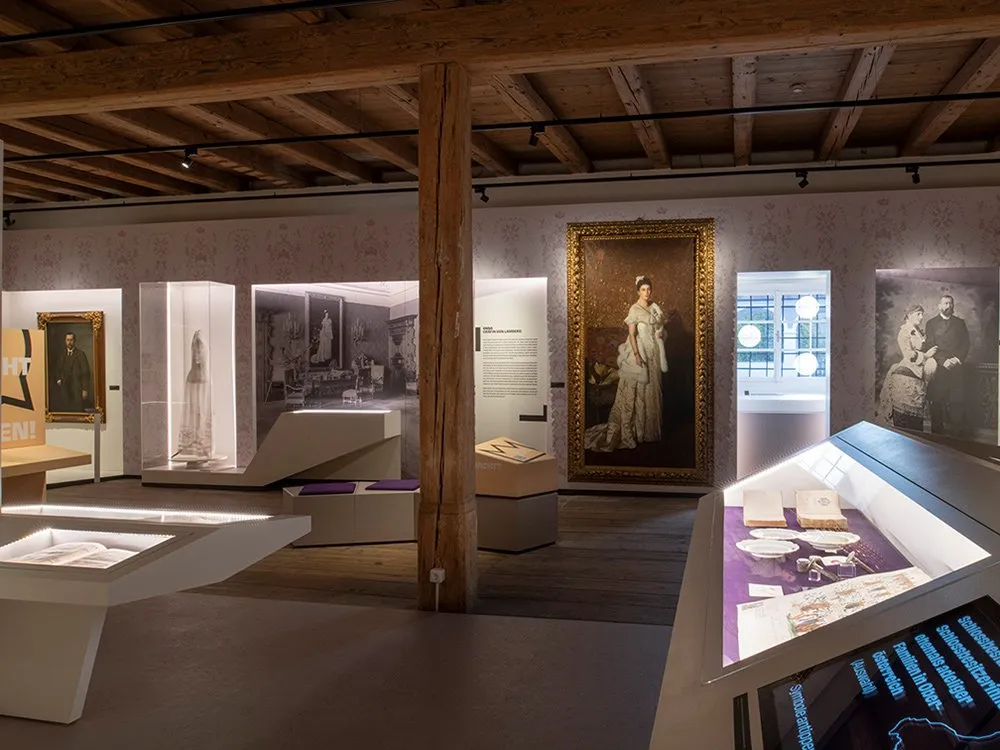 Ausstellungsraum: Vitrinen zeigen historische Kleidung und Porzellan. An den Wänden hängen Gemälde, darunter ein Porträt einer Frau in Weiß, sowie historische Fotos und Infotafeln.