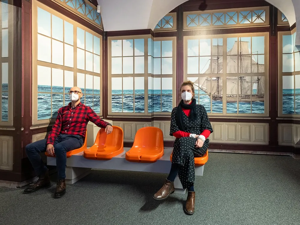 Ausstellungsraum: Zwei Personen sitzen auf einer Bank mit orangen Sitzen und betrachten Kunstwerke