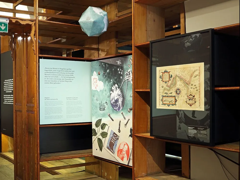 Ausstellungsraum: Holzregale mit einer Weltkugel, einer alten Karte und einem Bild mit Magellan-Themen, darunter Texte und Symbole wie Sterne und Schiffe.