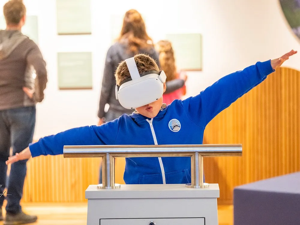 Ausstellungsraum: Kind mit VR-Brille steht an einer Konsole mit Metallstangen und breitet die Arme aus.