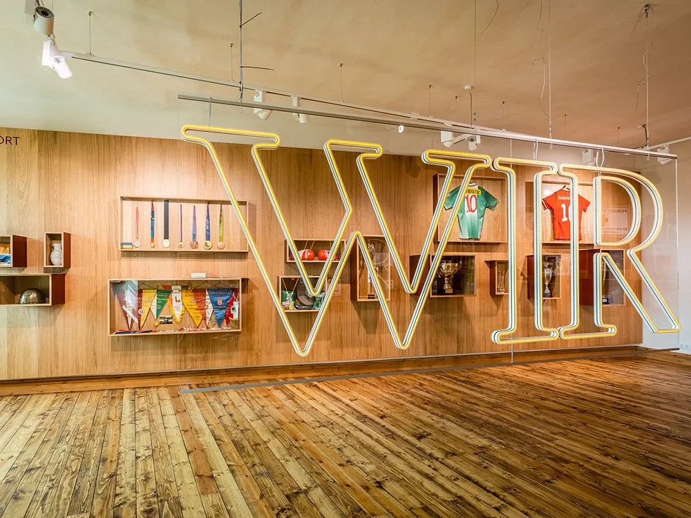 Helles Zimmer mit Holzwänden, beleuchteten Regalen mit Medaillen, Fahnen und Trophäen, sowie großen neonbeleuchteten Buchstaben „WIR“ und hängenden Sporttrikots.