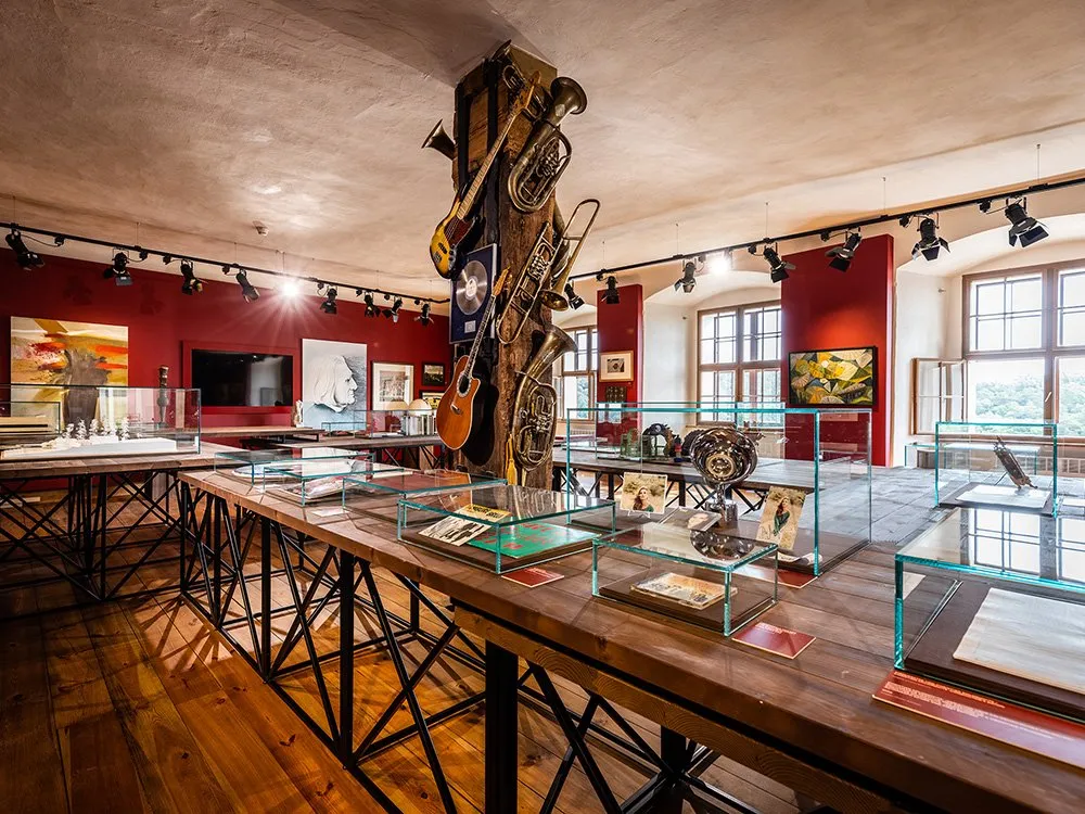 Ausstellungsraum mit Tischen mit Glasvitrinen, gefüllt mit Exponaten, sowie einer Säule mit hängenden Musikinstrumenten und Bildern.