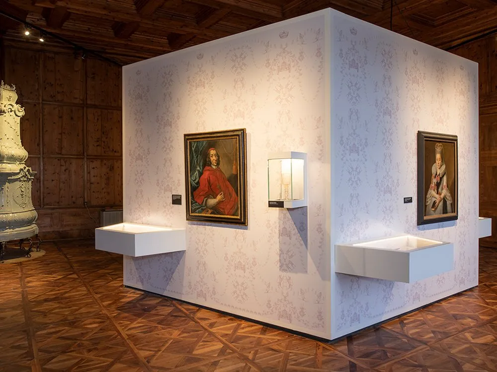 Ausstellungsraum: Ein quadratischer Raum mit floral gemusterter Tapete. An den Wänden hängen zwei Porträts, darunter eines eines Mannes in rotem Gewand. Vitrinen zeigen historische Objekte, ergänzt durch eine dekorative Kachelstube im Hintergrund.