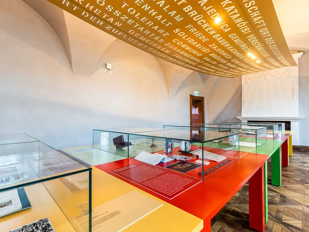 Ausstellungsraum mit bunt gestalteten Tischen mit Glasvitrinen und Exponaten, sowie einem überhängenden Banner mit Text.