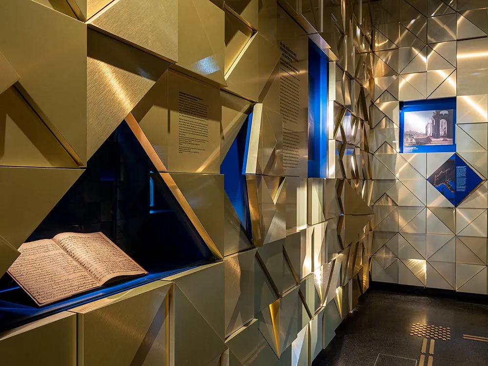 Detailansicht: Goldene geometrische Wand mit einer Vitrine und einem offenem Buch