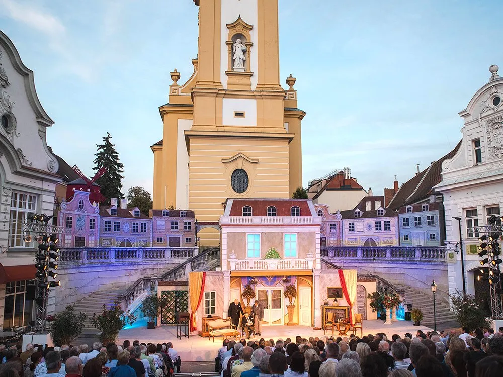 Bühnenbild vor einer Kirche. 3 Schauspielern auf der Bühne vor Publikum