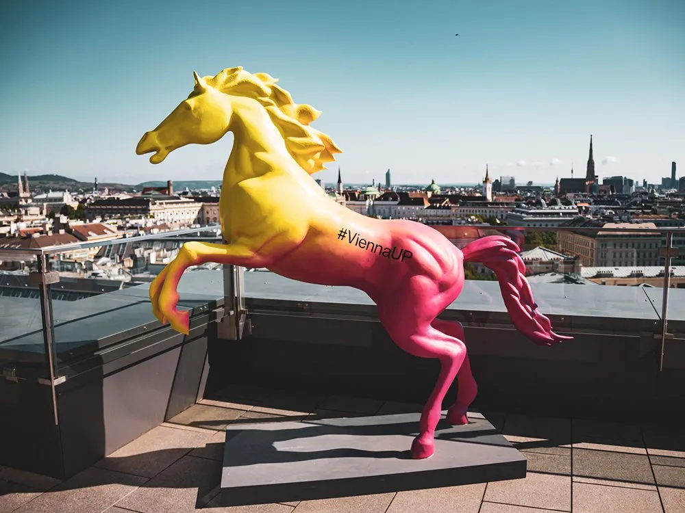 Dachterrassenansicht mit einer großen, zweifarbigen Pferdeskulptur in Gelb und Pink mit „#ViennaUP“, vor der Wiener Skyline.