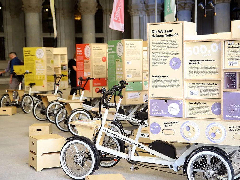 Weiße E-Lastenfahrräder mit Holzkisten und Plakaten zu Ernährung und Grünraum. Im Hintergrund Säulen und Fahnen mit „Wien macht gutes Klima“.