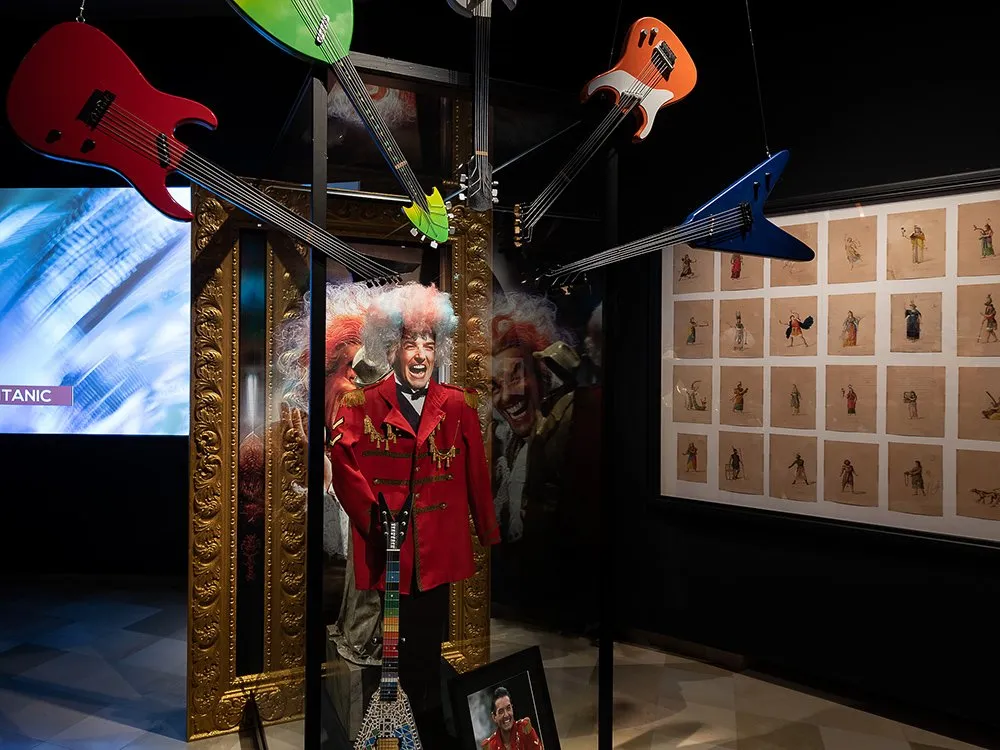 Ausstellungsraum: Falco-Figur im roten Mantel, umgeben von bunten Gitarren, die von der Decke hängen.